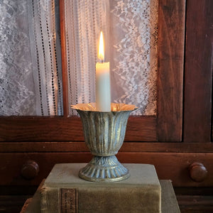Our Aged Gold Fluted Urn Taper Candle Cup features a distressed gold metal finish with an ornate scalloped design. The cup sits on a pedestal and has plenty of space between the holder and the sides, making it the perfect choice for displaying a candle ring! The candle cup has vintage style and is ideal for catching dripping wax. Measures 3.75" in diameter and 4"H and holds a standard taper (candle not included).