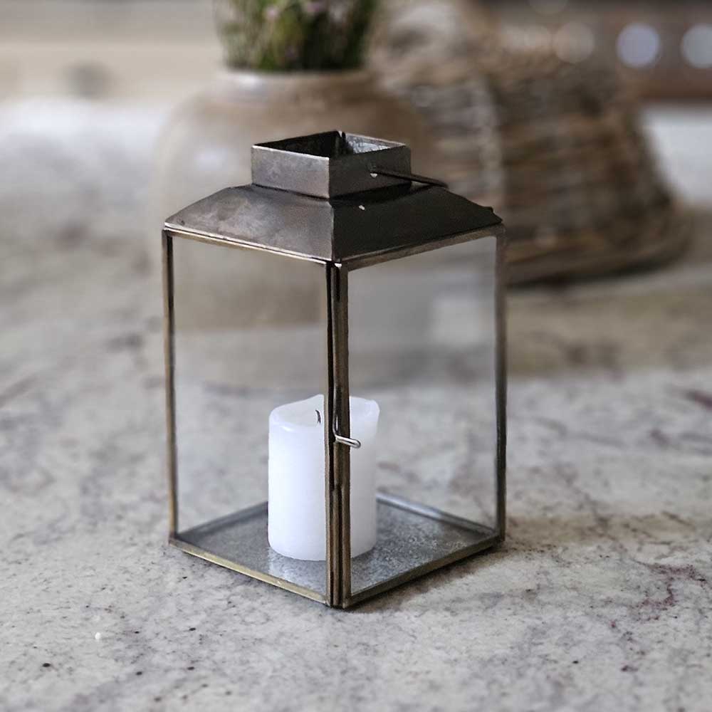 The Aged Iron Lantern adds rustic ambiance to any occasion.&nbsp; Features an antique bronze style finish and glass windows to protect the flame. Includes a door on one side and a wire handle for hanging. Makes a beautiful tabletop presentation. Holds one pillar candle. Each sold separately.