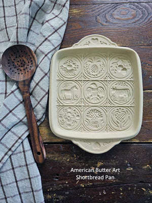 American Butter Art Shortbread Pan on a wooden surface with a wooden spoon and checkered cloth.