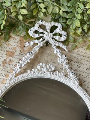 The Antoinette Bow Mirror is brimming with ornate details that add instant ambiance to any room. The distressed white mirror brings charming reflections with its lovely metal frame featuring an attractive bow and ribbon design. 7.75" x 12.75"H