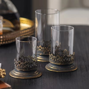 The Antiqued Gold and Glass Candle Hurricanes are an elegant set of three candle holders that features beautifully detailed metal bases with an antique black and gold finish. Perfect for adding warm ambiance and a touch of vintage charm to living rooms, dining tables, or event decor.