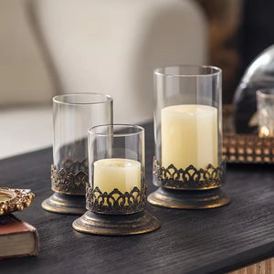 The Antiqued Gold and Glass Candle Hurricanes are an elegant set of three candle holders that features beautifully detailed metal bases with an antique black and gold finish. Perfect for adding warm ambiance and a touch of vintage charm to living rooms, dining tables, or event decor.