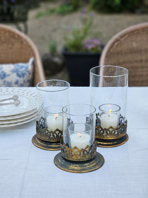 The Antiqued Gold and Glass Candle Hurricanes are an elegant set of three candle holders that features beautifully detailed metal bases with an antique black and gold finish. Perfect for adding warm ambiance and a touch of vintage charm to living rooms, dining tables, or event decor. Set of three.