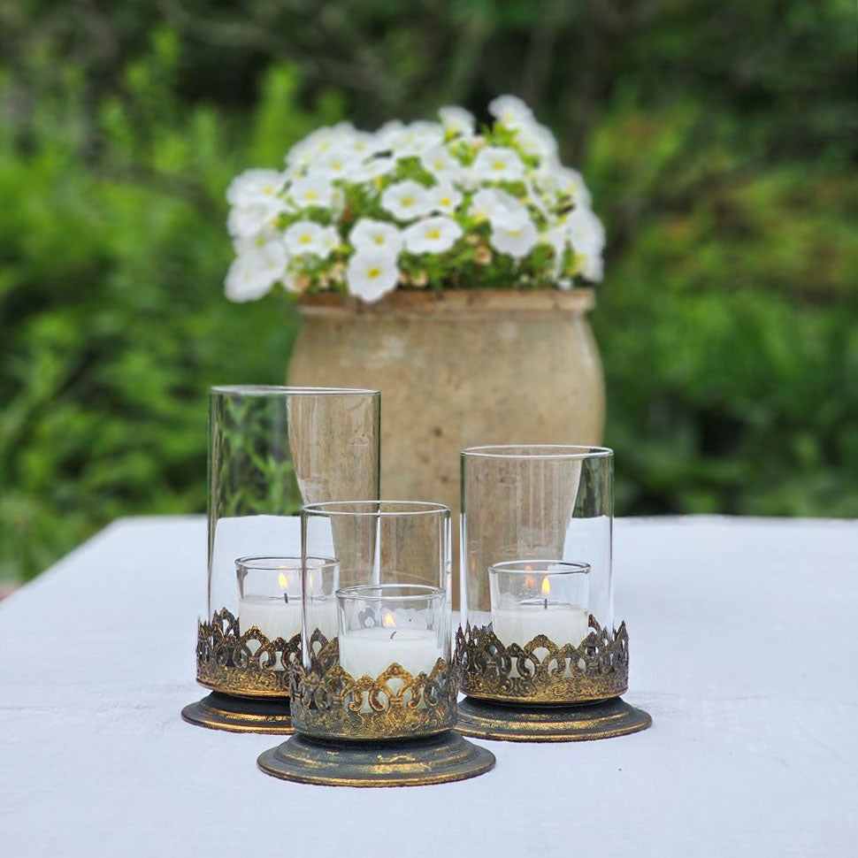 The Antiqued Gold and Glass Candle Hurricanes are an elegant set of three candle holders that features beautifully detailed metal bases with an antique black and gold finish. Perfect for adding warm ambiance and a touch of vintage charm to living rooms, dining tables, or event decor. Set of three.