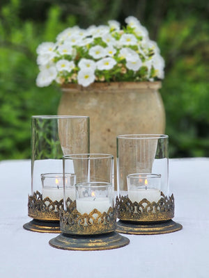 The Antiqued Gold and Glass Candle Hurricanes are an elegant set of three candle holders that features beautifully detailed metal bases with an antique black and gold finish. Perfect for adding warm ambiance and a touch of vintage charm to living rooms, dining tables, or event decor. Set of three.