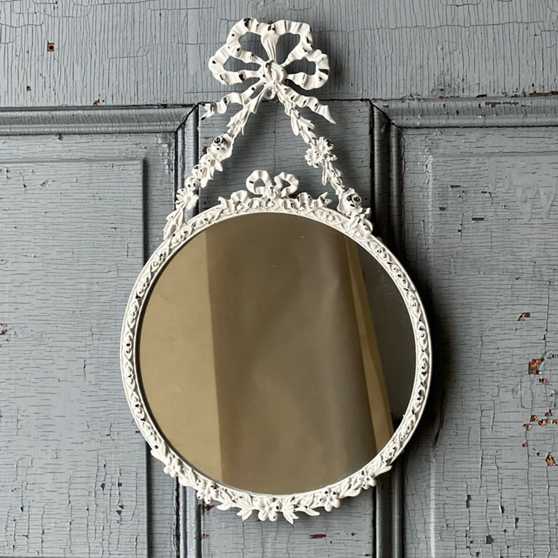 The Antoinette Bow Mirror is brimming with ornate details that add instant ambiance to any room. The distressed white mirror brings charming reflections with its lovely metal frame featuring an attractive bow and ribbon design. 7.75" x 12.75"H