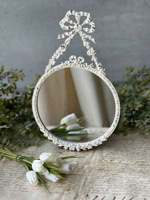 The Antoinette Bow Mirror is brimming with ornate details that add instant ambiance to any room. The distressed white mirror brings charming reflections with its lovely metal frame featuring an attractive bow and ribbon design. 7.75" x 12.75"H