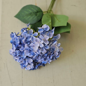 The Blue Hydrangea Bush Pick showcases exquisite blue fabric petals paired with fresh green leaves, creating a refined summer accent. Its flexible plastic stem supports bright hydrangea blooms subtly touched with soft white hues, adding depth and elegance. Measuring 17” long by 10” wide and 5” deep, this floral pick brings a sophisticated touch to any arrangement or decor.