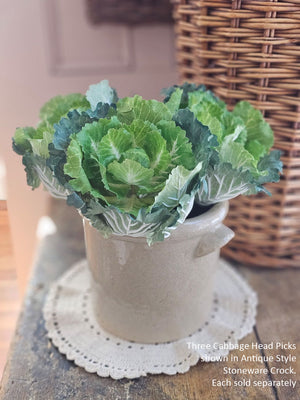 Artificial green cabbage plant in a stone crock on a wooden surface with wicker basket in the background.