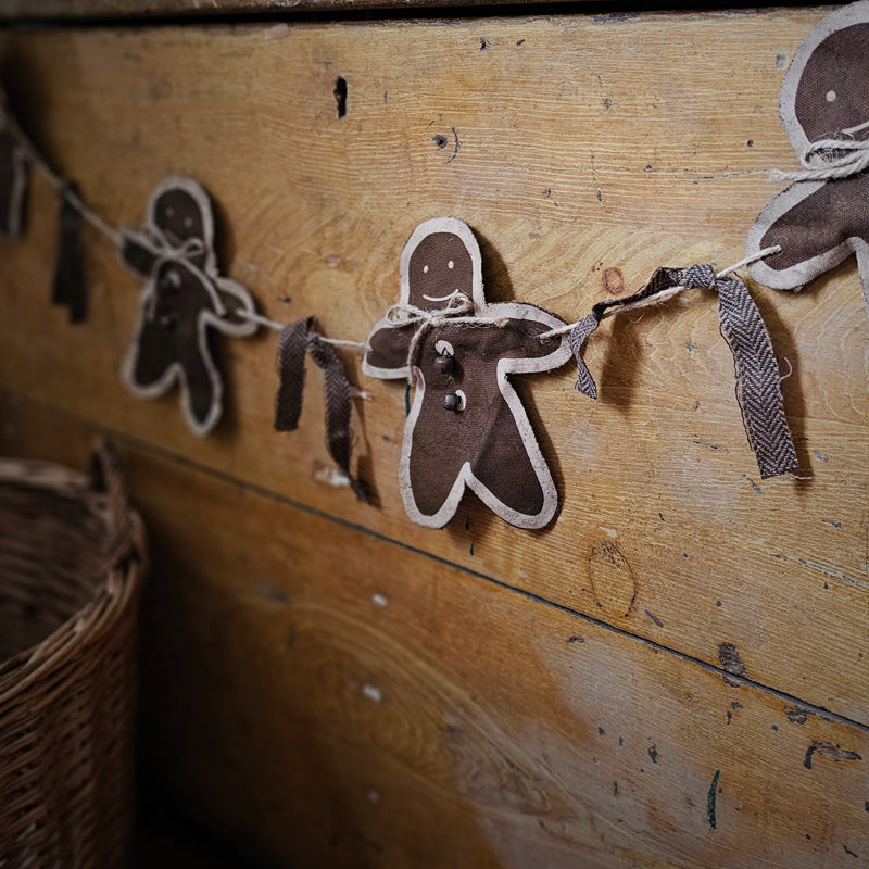 The fabric Gingerbread Garland with Primitive Style is a rustic Christmas garland made of brown fabric gingerbread man cutouts strung onto jute rope.  The gingerbread men have painted edges, jute rope bows, and rustic jingle bell buttons, and the garland is decorated with brown herringbone ribbon. Both ends of the garland include jute rope loops to use for display, whether across a banister, mantle, doorway, window, or wall. Feature four gingerbread man cutouts. Measures 7” high by 36” long.