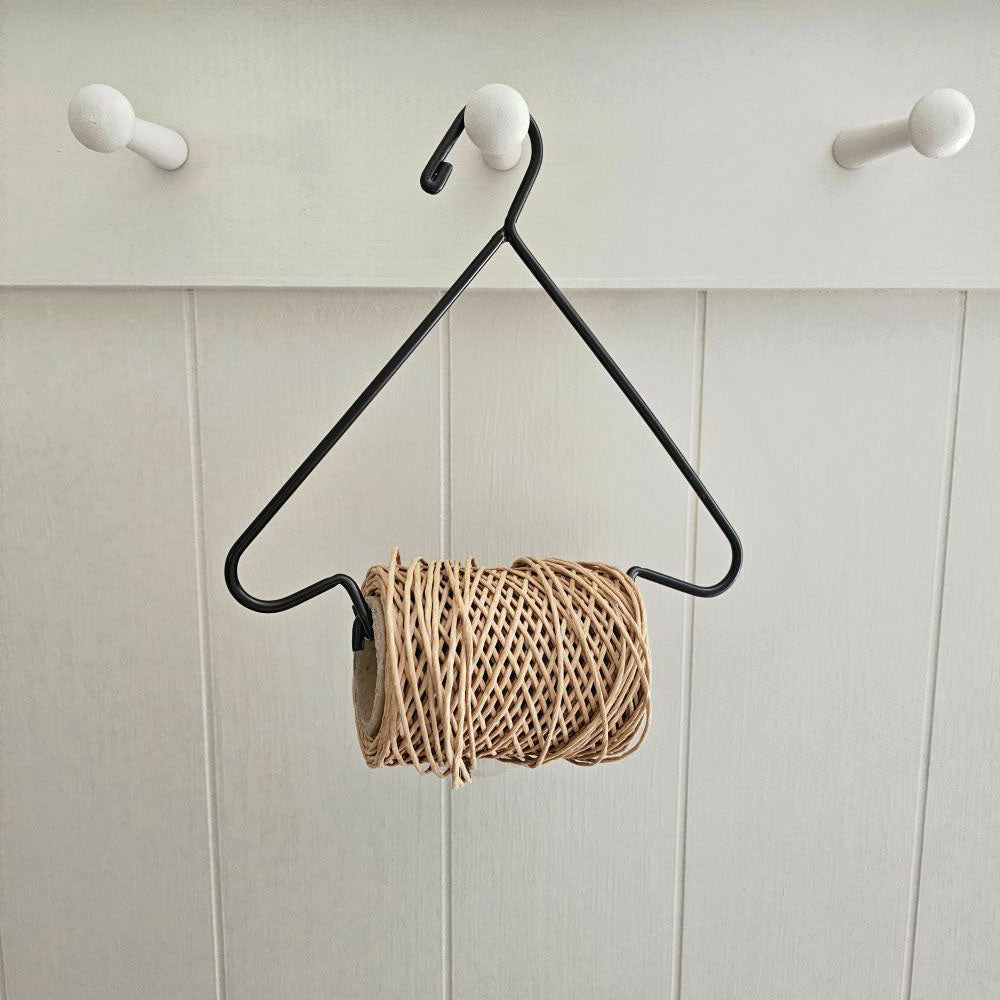 A black metal hanger with a triangular frame and a roll of natural jute twine wrapped around it, hanging against a white paneled wall with three white pegs.