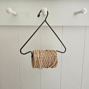 A black metal hanger with a triangular frame and a roll of natural jute twine wrapped around it, hanging against a white paneled wall with three white pegs.