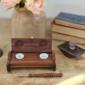 The Inkwell Stand with Porcelain Inkwells and Wood Nib Pen Set makes a perfect desk accessory reminiscent of early Colonial days. The set features a wood inkwell stand that opens and closes, two white porcelain inkwells, a wood nib writing pen, and a bag black ink powder so you can easily get started.