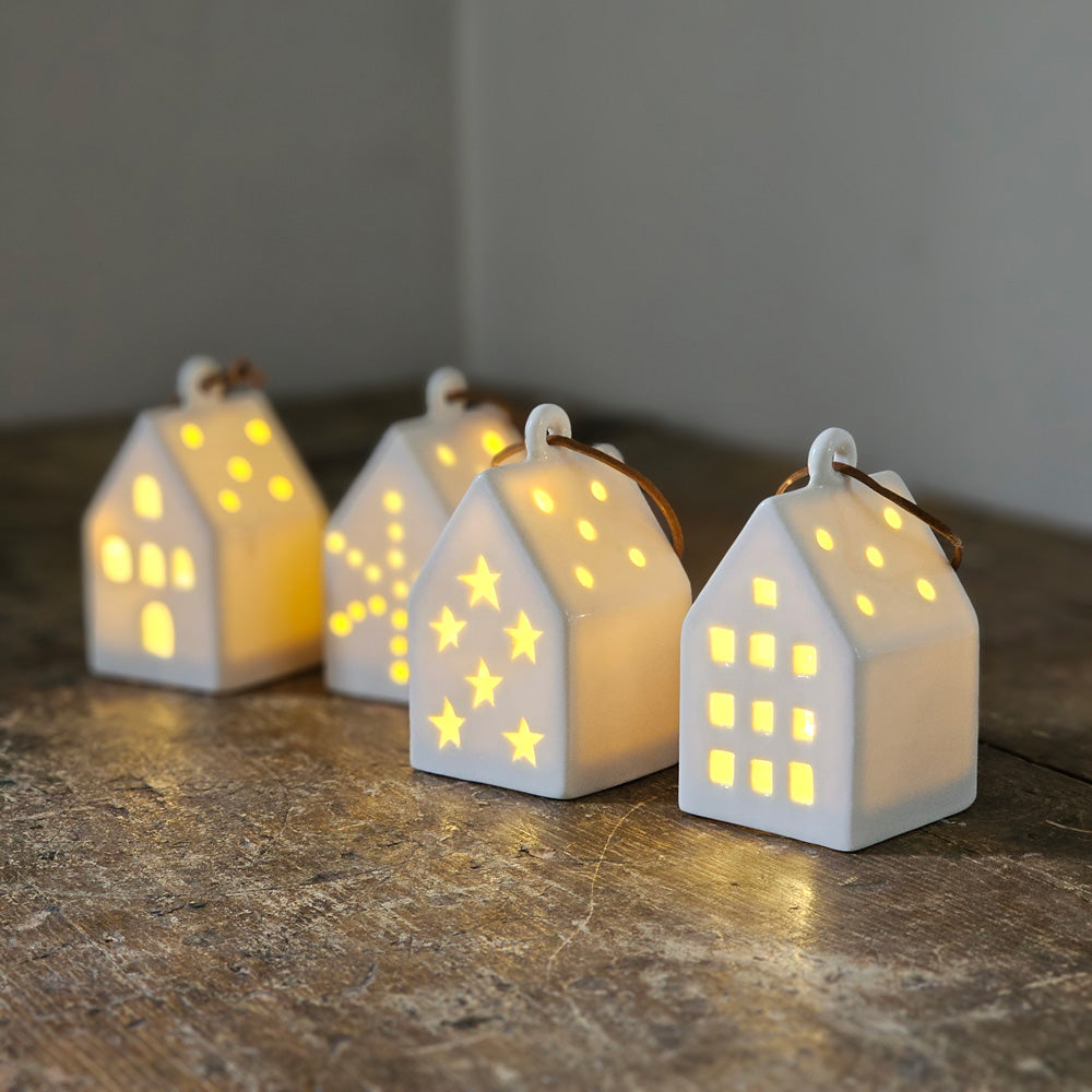 Create magical displays or add warmth to your Christmas tree with this Lighted Ceramic House Ornament Set. The set of four has a quiet elegance with an organic, minimalist design. The little white ceramic houses look beautiful even when they are glowing with the warm LED light. Each has a leather strap for hanging. Each has a different style. Perfect for traditional or modern farmhouse decor. Requires two LR44 batteries (included). Set of four. Each is 2"L x 2"W x 3.5"H