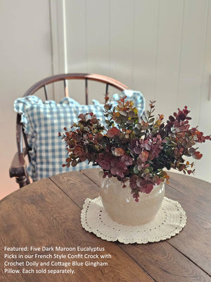 Bouquet of dark maroon eucalyptus in a French-style confit crock on a wooden table with a checkered pillow in the background.