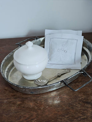 White ceramic lidded container on a metal tray with a spoon and lace napkin on a wooden surface.