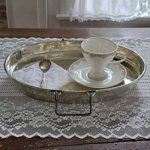 A vintage silver metal tea tray with side handles, displayed on a wooden table over a white lace runner. The tray is styled with a white teacup and saucer featuring gold trim, a white napkin, and a silver spoon.