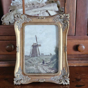 Ornate Gold Frame with Vintage Windmill Print adds old-world charm to any room. The aged gold frame can be used with or without the print and can hang on a wall or sit on a tabletop, both vertically or horizontally. Made of resin, the frame features a wood-like feel with ornately detailed edging. Includes glass and fits 5 x 7 prints. The print features a moody landscape with cloudy skies with a windmill and a woman carrying her grain sack to mill. Overall size is: 7.5" x 9.5"