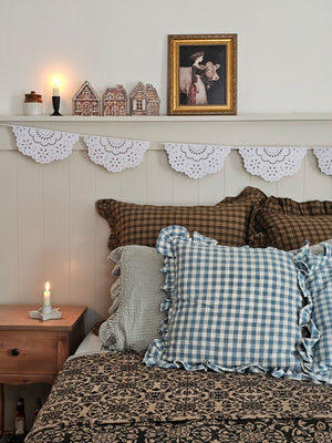 Cozy bedroom with checkered and plaid pillows, wooden nightstand, and decorative shelf with gingerbread houses and milkmaid print.