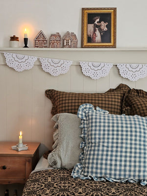 Cozy bedroom with checkered and plaid pillows, wooden nightstand, and decorative shelf with gingerbread houses and milkmaid print.
