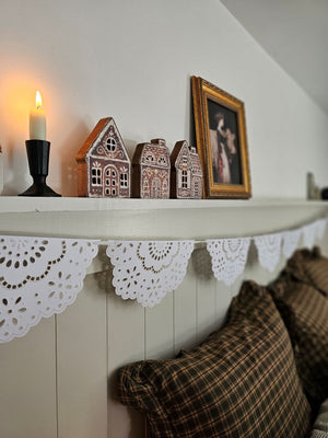 Cozy bedroom with checkered and plaid pillows, wooden nightstand, and decorative shelf with gingerbread houses and milkmaid print.