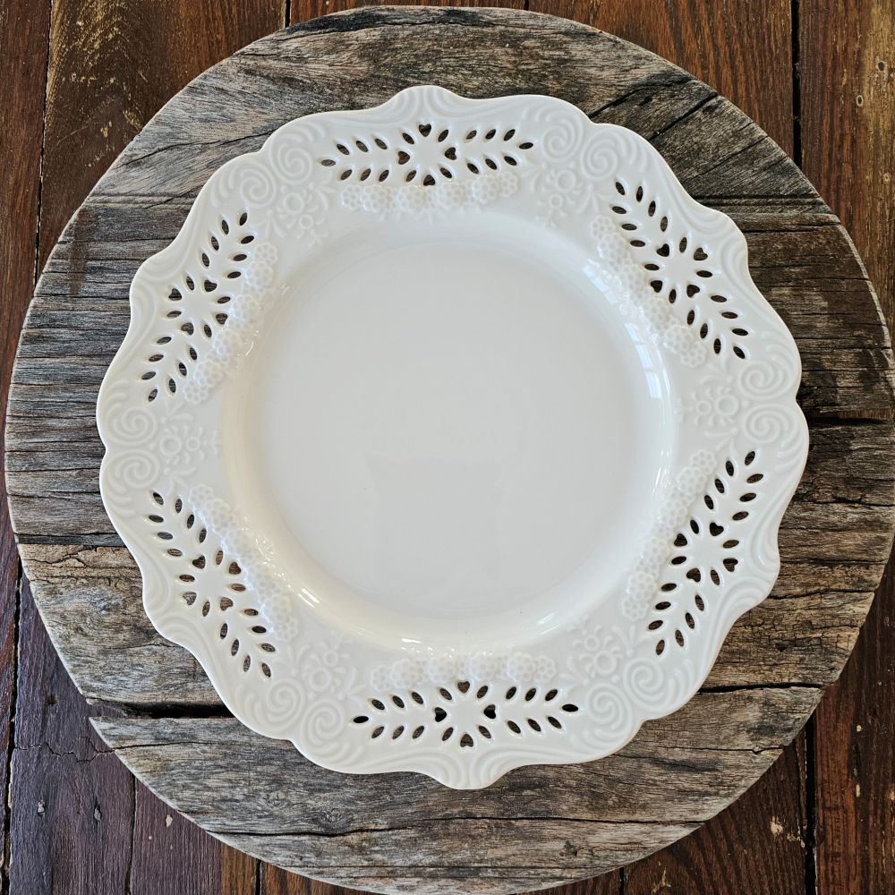 This creamy white porcelain dinner plate marries vintage charm with refined elegance. Featuring hand-detailed embossed flowers and delicate cutout lace work, the gracefully scalloped edge evokes timeless sophistication. Whether gracing your everyday table or reserved for special gatherings, this single plate serves as a subtle statement of cultivated taste and enduring style. Food safe. Microwave safe. Hand wash recommended. Each sold separately. 10" Diam