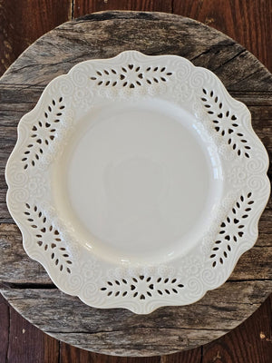 This creamy white porcelain dinner plate marries vintage charm with refined elegance. Featuring hand-detailed embossed flowers and delicate cutout lace work, the gracefully scalloped edge evokes timeless sophistication. Whether gracing your everyday table or reserved for special gatherings, this single plate serves as a subtle statement of cultivated taste and enduring style. Food safe. Microwave safe. Hand wash recommended. Each sold separately. 10" Diam