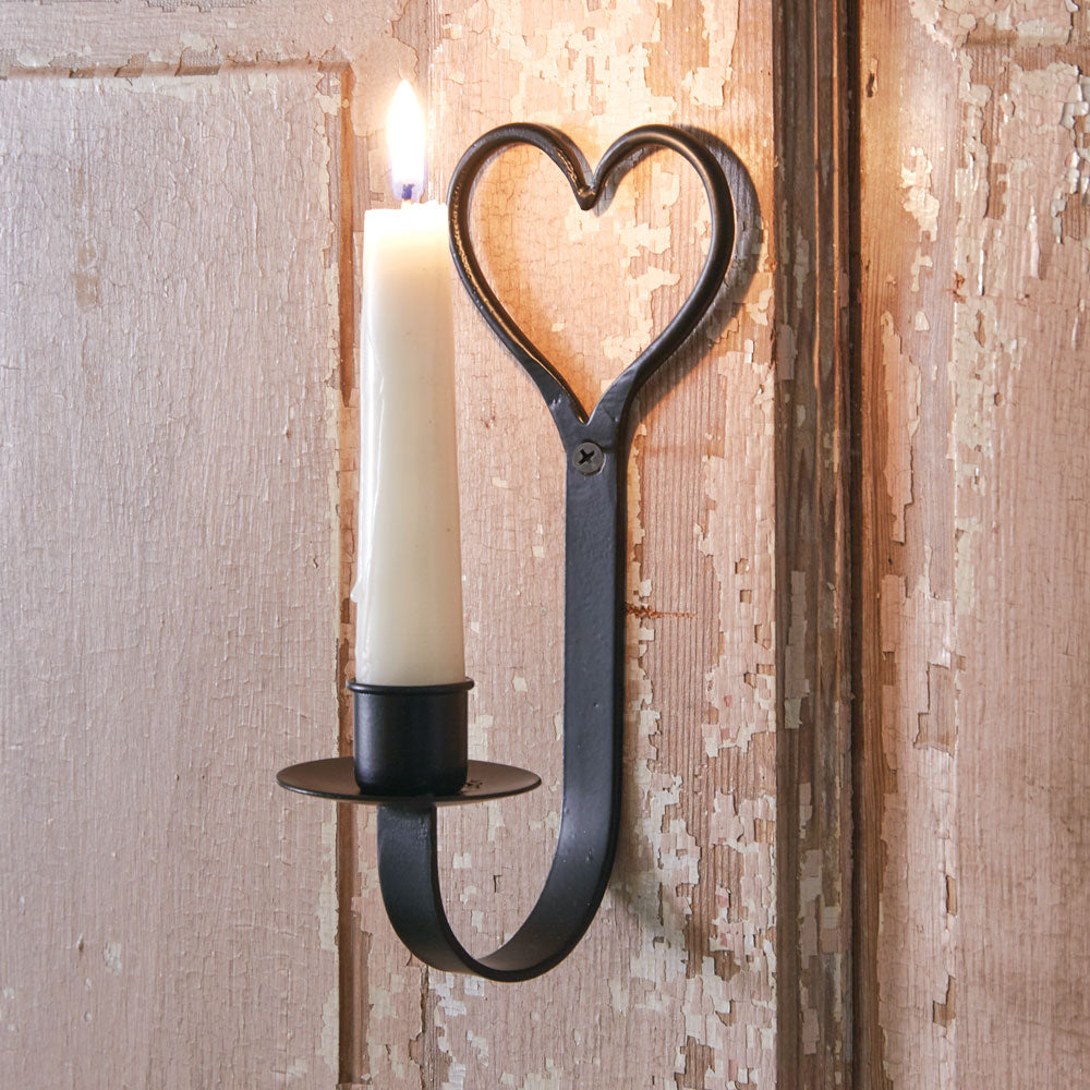 The Primitive Heart Taper Candle Sconce adds a sweet touch to any room. The relaxed design features a metal taper holder with black finish. The top of the holder features a beautiful heart shape and a pre-drilled hole for hanging. The holder curves up at the base and holds a candle tray with a taper candle cup. 3''W x 3½''D x 7¼''H