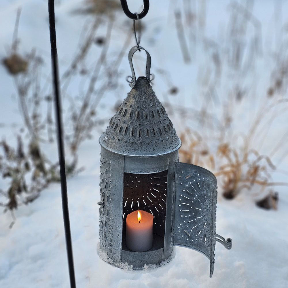 This authentic Colonial reproduction captures the rustic charm of a vintage watchman's lantern with meticulous attention to detail. Crafted from solid metal with a weathered zinc patina, it accommodates a standard 3-inch pillar candle and features an integrated round hook for suspended display or wall mounting. The punched tin design creates a magical glow. 