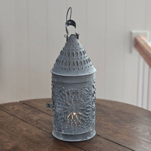 This authentic Colonial reproduction captures the rustic charm of a vintage watchman's lantern with meticulous attention to detail. Crafted from solid metal with a weathered zinc patina, it accommodates a standard 3-inch pillar candle and features an integrated round hook for suspended display or wall mounting. The punched tin design creates a magical glow. 