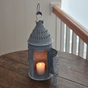 This authentic Colonial reproduction captures the rustic charm of a vintage watchman's lantern with meticulous attention to detail. Crafted from solid metal with a weathered zinc patina, it accommodates a standard 3-inch pillar candle and features an integrated round hook for suspended display or wall mounting. The punched tin design creates a magical glow. 