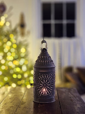 This authentic Colonial reproduction captures the rustic charm of a vintage watchman's lantern with meticulous attention to detail. Crafted from solid metal with a weathered zinc patina, it accommodates a standard 3-inch pillar candle and features an integrated round hook for suspended display or wall mounting. The punched tin design creates a magical glow. 