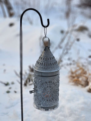 This authentic Colonial reproduction captures the rustic charm of a vintage watchman's lantern with meticulous attention to detail. Crafted from solid metal with a weathered zinc patina, it accommodates a standard 3-inch pillar candle and features an integrated round hook for suspended display or wall mounting. The punched tin design creates a magical glow. 