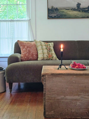 A cozy living room scene featuring a dark gray fabric sofa with two decorative throw pillows, a rustic wooden coffee table with a Primitive Blacksmith Taper Candle Holder and a plate of red grapes, and a window with white curtains in the background.