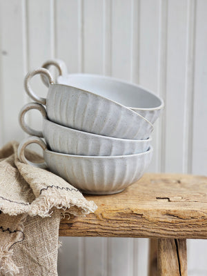 Rustic Ceramic White Bowl with handle keeps a low profile, making it perfect for coffee or soup. Add a candle and it turns into a beautiful chamberstick. This stoneware mug features a white finish speckled with clay tones peaking through the ribbed design. It brings an earthy elegance to your farmhouse kitchen. Each sold separately. Includes one bowl with handle.