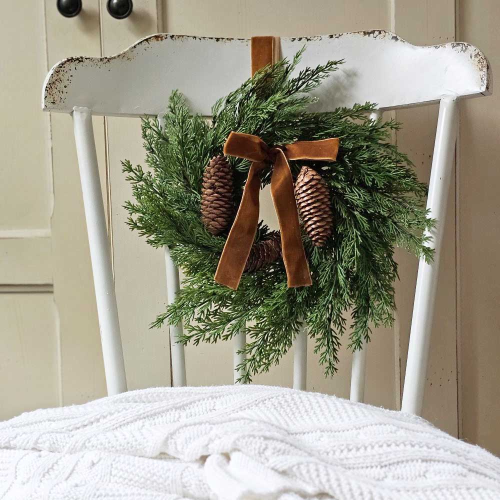 The Small Cedar Wreath with Velvet Bow and Hanging Ribbon adds a refined touch to kitchen cabinets, windows, and cupboards. Crafted with realistic faux cedar foliage and pinecones, it brings understated elegance to any space. Finished with a plush velvet bow and an attached ribbon, this wreath offers effortless and graceful hanging. 13" Diam