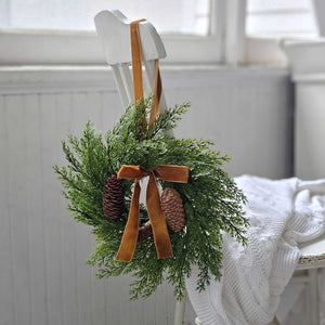 The Small Cedar Wreath with Velvet Bow and Hanging Ribbon adds a refined touch to kitchen cabinets, windows, and cupboards. Crafted with realistic faux cedar foliage and pinecones, it brings understated elegance to any space. Finished with a plush velvet bow and an attached ribbon, this wreath offers effortless and graceful hanging. 13" Diam