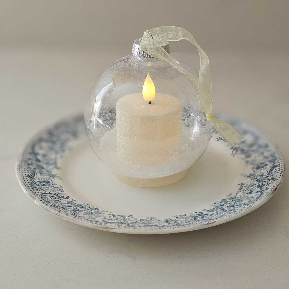 The lighted Snow Globe Candle Ornament lends a quiet elegance with a minimalist feel. The clear ornament bulb contains a battery operated white votive candle that sits on a dusting of sparkling faux snow. The LED candle has a subtle flicker for added ambiance. A flat base allows for freestanding display on a surface, and a light gold ribbon allows for hanging display. The bottom features a switch that can be used to set a timer or for easy on-and-off. The candle requires 1 CR2032 battery, which is included.