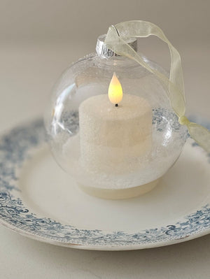 The lighted Snow Globe Candle Ornament lends a quiet elegance with a minimalist feel. The clear ornament bulb contains a battery operated white votive candle that sits on a dusting of sparkling faux snow. The LED candle has a subtle flicker for added ambiance. A flat base allows for freestanding display on a surface, and a light gold ribbon allows for hanging display. The bottom features a switch that can be used to set a timer or for easy on-and-off. The candle requires 1 CR2032 battery, which is included.