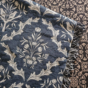 Blue and white floral and thistle patterned throw blanket with black tassels on a wooden surface. Made in the USA