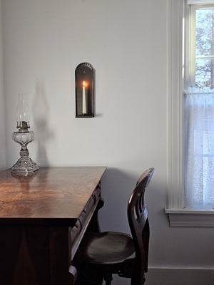 Our Colonial House Taper Candle Wall Sconce brings historic ambiance to any room. Made of tin, with a gunmetal grey finish, this wall sconce features a decorative fluted edge around the top of the piece. Its simple design works well with rustic farmhouse,  coastal cottage, or Early American decor. Holds one regular taper candles (not included). Made in the USA.  4.5”L x 3.5”W(depth) x 14”H