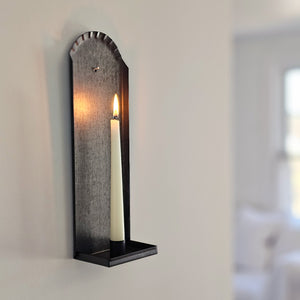 Our Colonial House Taper Candle Wall Sconce brings historic ambiance to any room. Made of tin, with a gunmetal grey finish, this wall sconce features a decorative fluted edge around the top of the piece. Its simple design works well with rustic farmhouse,  coastal cottage, or Early American decor. Holds one regular taper candles (not included). Made in the USA.  4.5”L x 3.5”W(depth) x 14”H