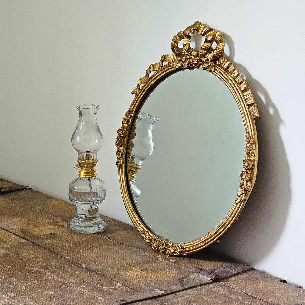 This Vintage Style Gold Mirror brings Gilded Age ambiance to any room. Made of pewter, this round mirror has an aged gold finish for a luxurious look. Embellished with faux diamonds and a golden bow, the ornate design will add a touch of sophistication and antique style charm to your home. 11.8"x1.2"D x13.6"H