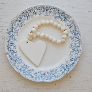 This White Clay Heart and Wood Bead Ornament adds texture and style to hooks and shelves. The ornament features natural wood beads and a simple white clay heart dangling on the end. Perfect for wood and white farmhouse decor. 2.75" W x 7.5"H