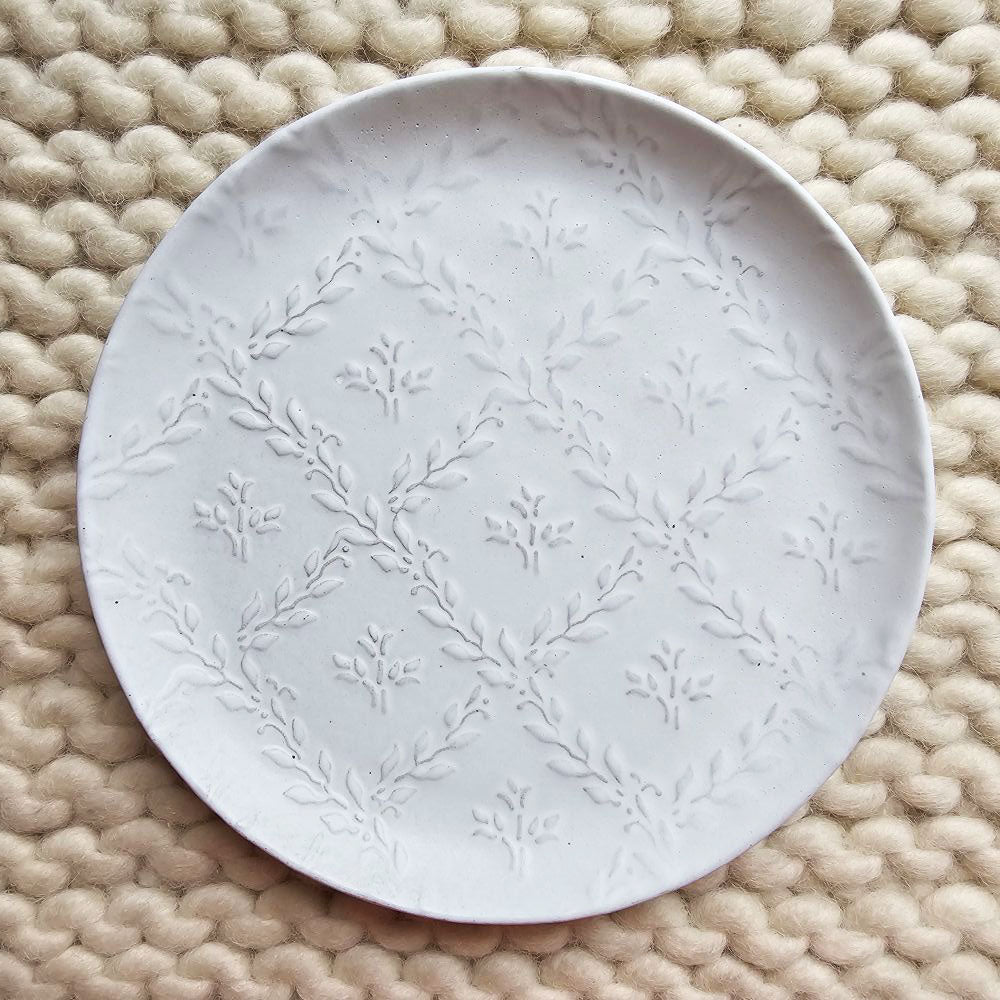 Add cheerful country style with our White Ceramic Embossed Plate. Made of ceramic, this durable plate displays a white glaze finish with embossed patterns of plant vines on the inside of the plate. Embossed vine designs come through the glaze for textured detail. Perfect for holding trinkets and jewelry or use as a soap dish. Includes one plate. Food safe. Hand wash. Each sold separately. Hand wash. 6" Diam