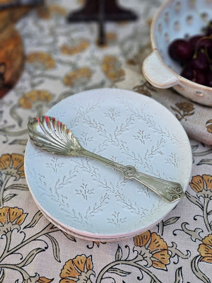 Decorative ceramic plate with embossed design and a silver spoon on a floral-patterned tablecloth.