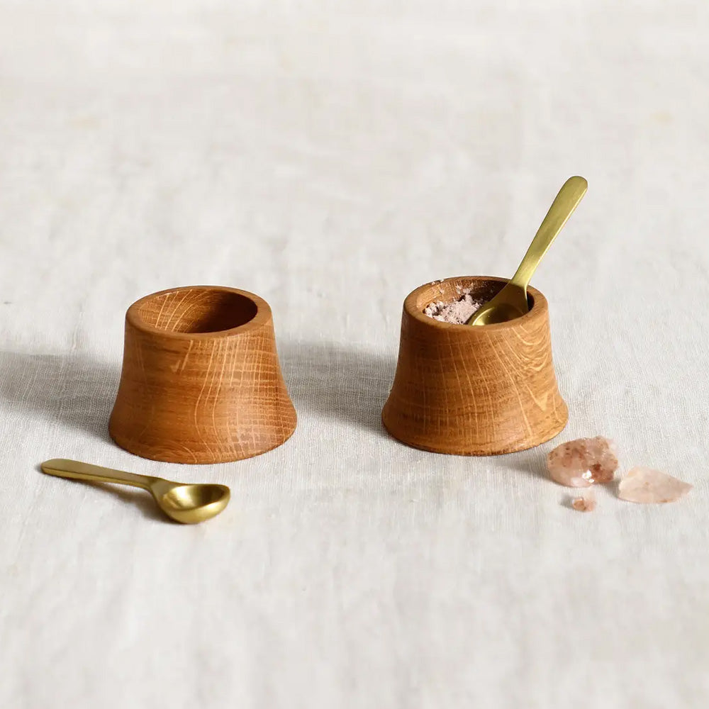 Crafted from premium French oak, this Wood Pinch Bowl and Spoon Set features hand-shaped tower-style bowls, perfect for presenting salt, pepper, or other condiments with elegance. The set includes two petite matte brass spoons, enhancing its refined appeal. Finished with a food-safe oil, each piece is designed for delicate hand washing, ensuring lasting beauty and function. Ideal for sophisticated table settings, these pinch bowls combine artisanal craftsmanship with practical usability. 