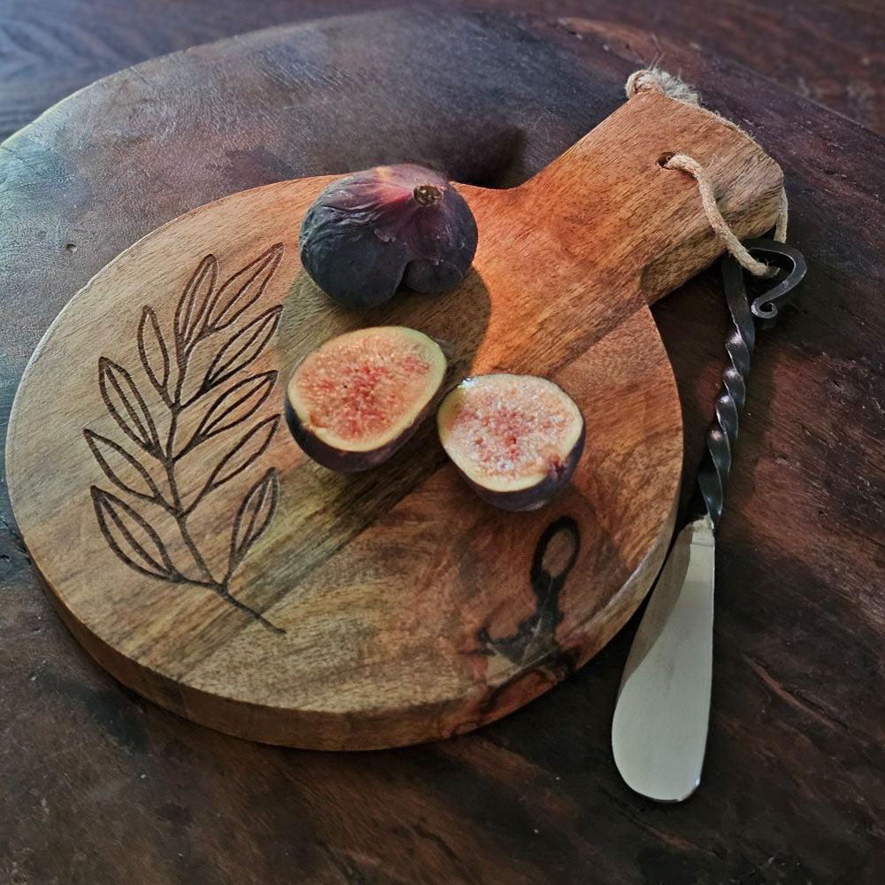 Wooden cutting board with engraved leaf design, sliced figs, and a knife on a dark surface.