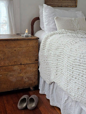 Close-up of a textured white pillow on a bed with wooden furniture and slippers in the foreground.