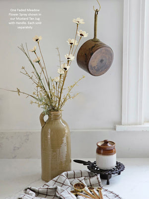 A rustic mustard tan jug vase holding Faded Meadow Flower Spray, styled on a white countertop with a small white and brown jar on a black stand, gold-toned utensils on a checkered cloth, and a brown hanging copper pot against a white wall.